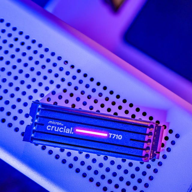  A Crucial T710 NVMe M.2 SSD with heatsink (indicator light on) resting on a white surface under pink and blue light.