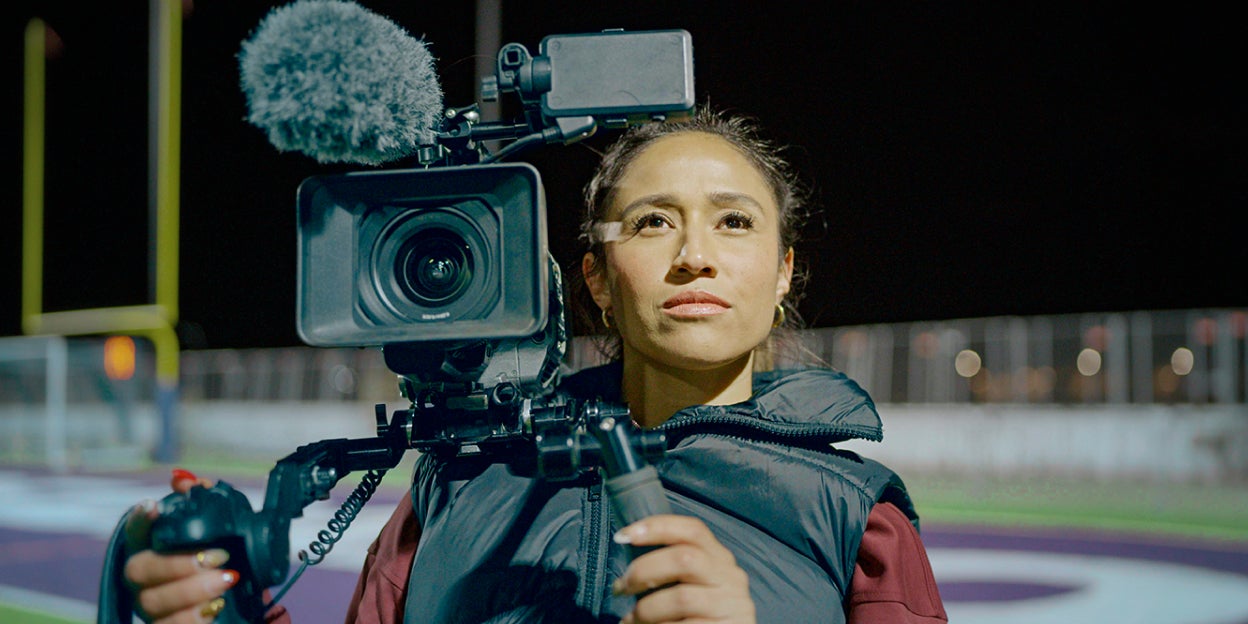Sarina Soriano standing on a football field and staring into the distance while holding a professional video camera.