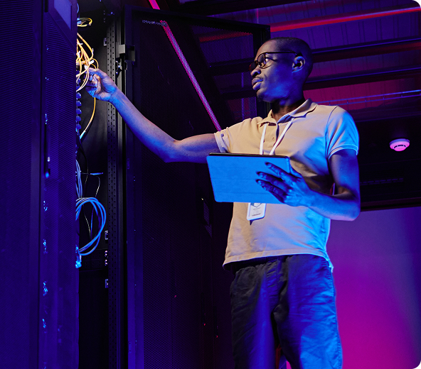 An IT technician holding a tablet examines a server rack and cables in a server room with pink and blue lighting.