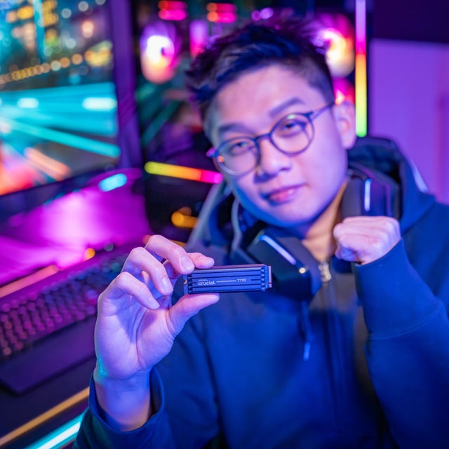 Gamer with glasses and headphones turned away from their RGB gaming PC and holding up a Crucial T710 to the camera.